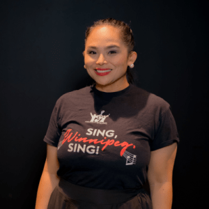 A woman smiles at the camera, standing against a dark background. She is wearing a black T-shirt that says Sing, Winnipeg, Sing! in white and red letters. She has pearl earrings and red lipstick.