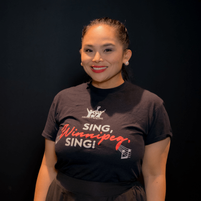 A woman smiles at the camera, standing against a dark background. She is wearing a black T-shirt that says Sing, Winnipeg, Sing! in white and red letters. She has pearl earrings and red lipstick.