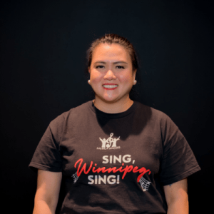 A woman with long dark hair tied back smiles at the camera, wearing a black t-shirt that reads SING, Winnipeg, SING! on a plain black background.