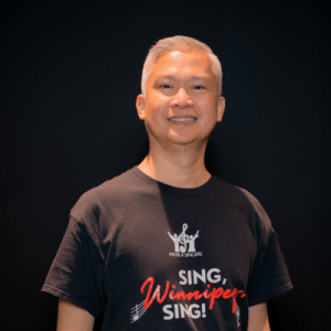 A smiling person with short gray hair stands in front of a black background, wearing a black t-shirt that reads SING, Winnipeg, SING! in white and red letters, with a musical logo above the text.