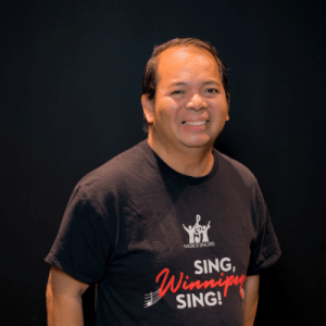 A smiling man with short dark hair stands against a plain black background, wearing a black t-shirt that reads Sing, Winnipeg, Sing! in white and red letters.