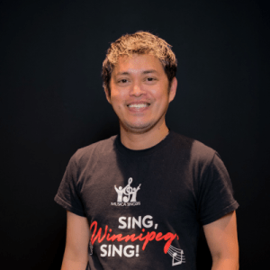 A smiling person with short, light brown hair stands in front of a black background, wearing a black T-shirt that reads SING, Winnipeg, SING! with a music note and MUSICA SINGERS logo.