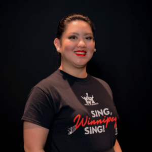 A woman with pulled-back hair and red lipstick smiles at the camera. She is wearing a black Sing, Winnipeg, Sing! t-shirt with pearl earrings, standing against a plain black background.