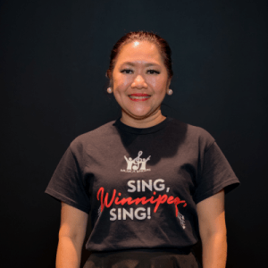 A woman with dark hair pulled back smiles at the camera. She is wearing a black t-shirt that says SING, Winnipeg, SING! on a plain dark background.