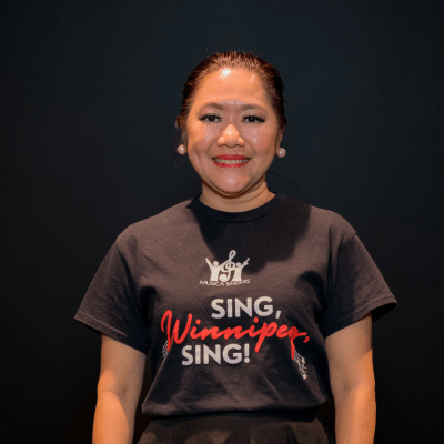 A woman with dark hair pulled back smiles at the camera. She is wearing a black t-shirt that says SING, Winnipeg, SING! on a plain dark background.