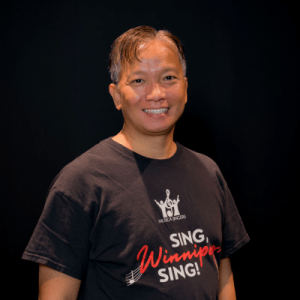 A person with short hair smiles at the camera, wearing a black T-shirt that reads MUSAIC SINGS SING, Winnipeg, SING! The background is black, highlighting the individual.
