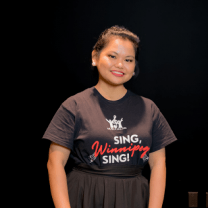 A woman with tied-back hair smiles while wearing a black Sing, Winnipeg, Sing! t-shirt, standing against a dark background.