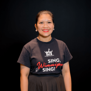 A woman with dark hair tied back, wearing pearl earrings and a black Musica Singers - Sing, Winnipeg, Sing! t-shirt, stands and smiles in front of a plain black background.
